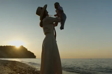 Woman lifting a baby in the air on a beach with the sun setting in the background.